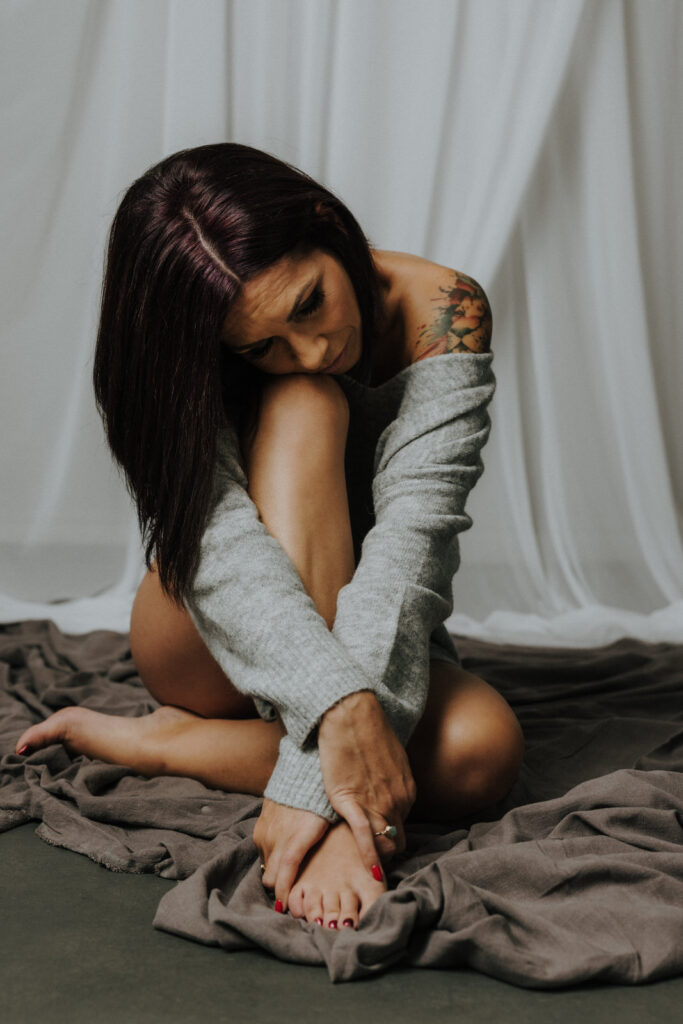boudoir image of woman sitting on floor on fabric wearing off shoulder sweater hugging legs
