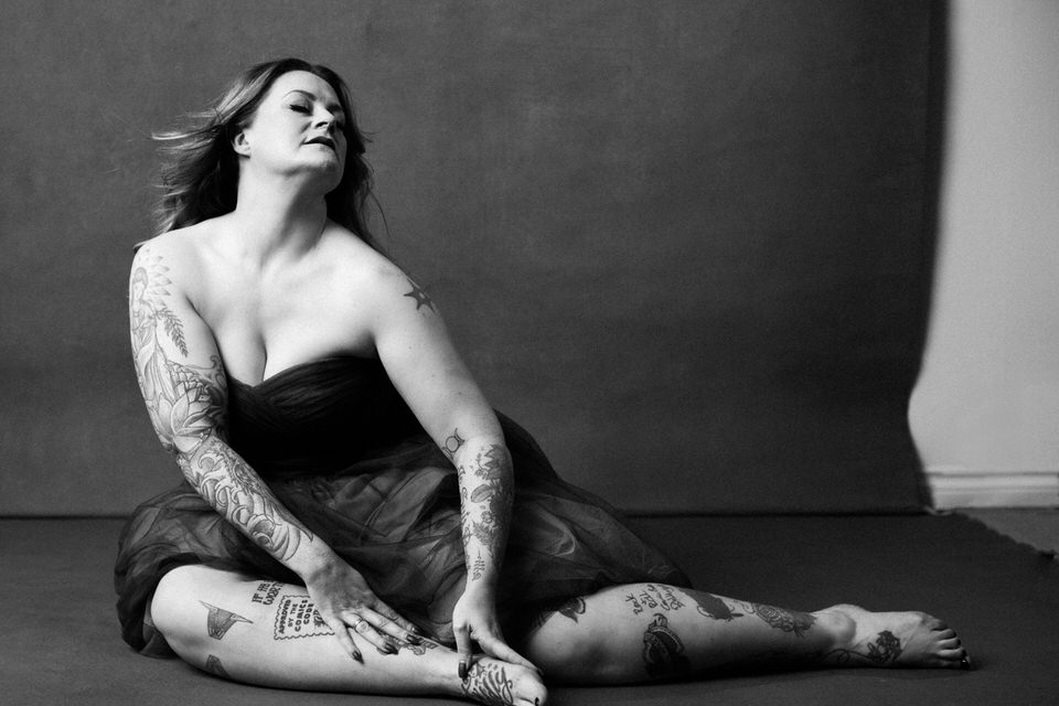 Black and white boudoir image of a tattooed woman wearing a strapless gown sitting on the floor and tilting her head backwards