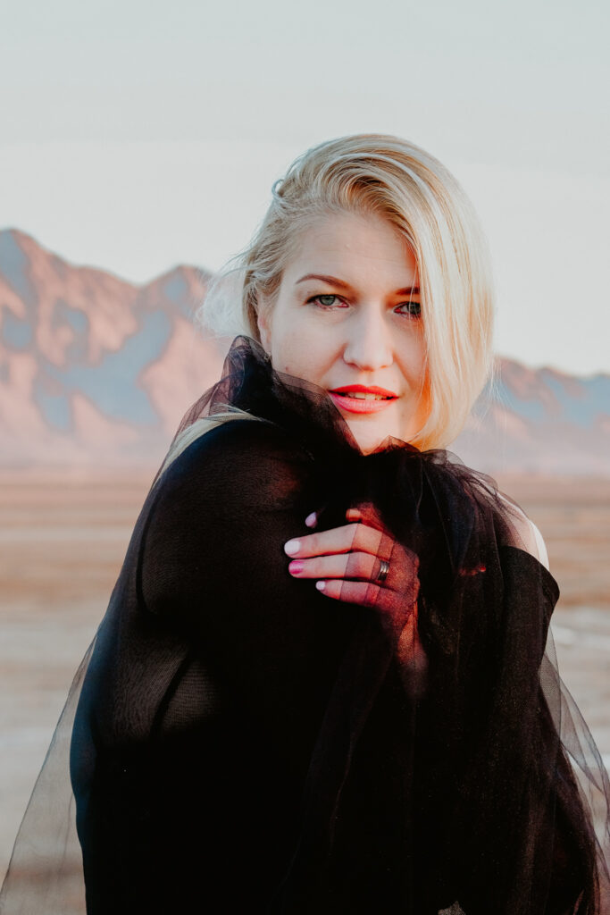 Woman with blonde hair wearing black and wrapped in black fabric with desert mountains in the background during a spontaneous boudoir shoot