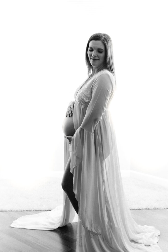 pregnant woman standing holding her bare belly wearing a white draped gown with belly showing