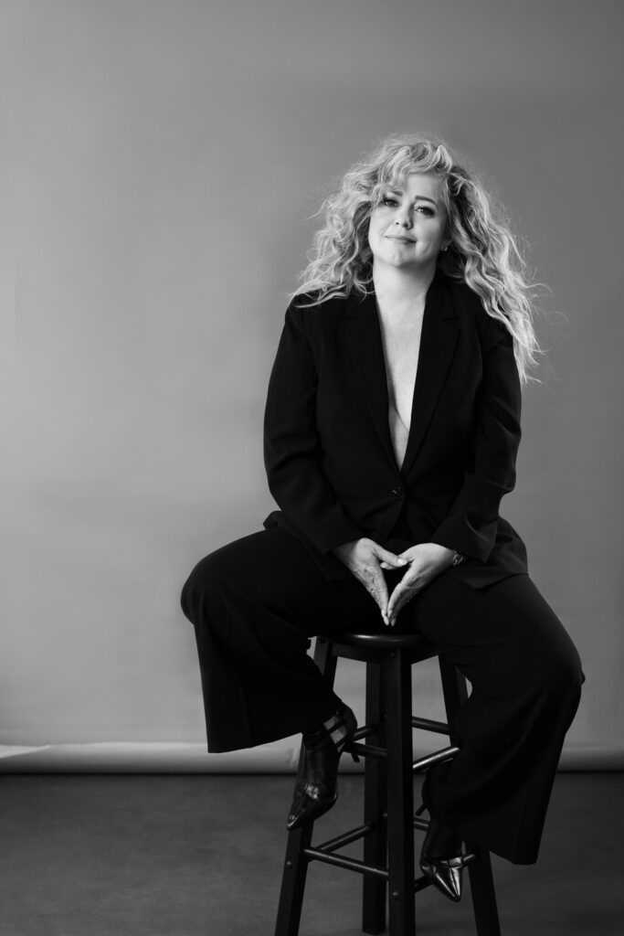 a luxe intimate portrait of a woman sitting on a stool with feet up on the rungs, she's wearing a cleavage baring power suit and her hands together in front of her