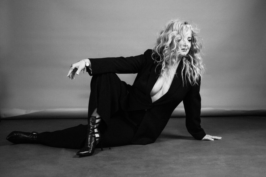 an intimate luxe portrait of a woman in a power suit showing cleavage, sitting on the floor with one leg stretched out and the other crossed up and over the other showing her strappy boots and one hand holding her up and the other dangling over her raised knee