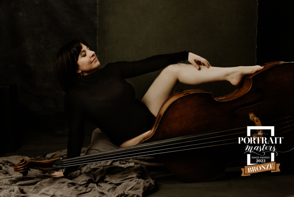 boudoir image of a woman in a black bodysuit sitting beside a laid down bass with one foot sitting atop the bass