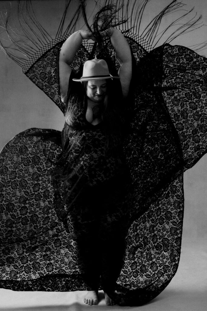 black and white boudoir image of woman wearing lacey robe and hat and arms in the air