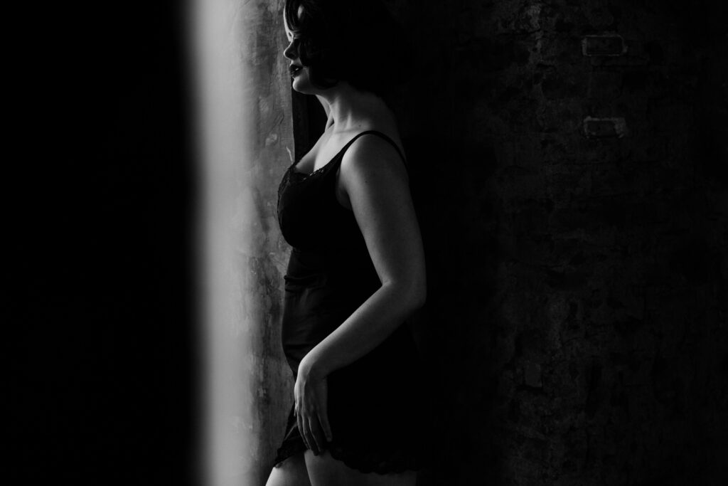 Black and white boudoir portrait of an white woman with a black slip dress leaning against a brick wall, the photo shows the side profile of her body created by Michele Mateus Vancouver Portrait Photograph