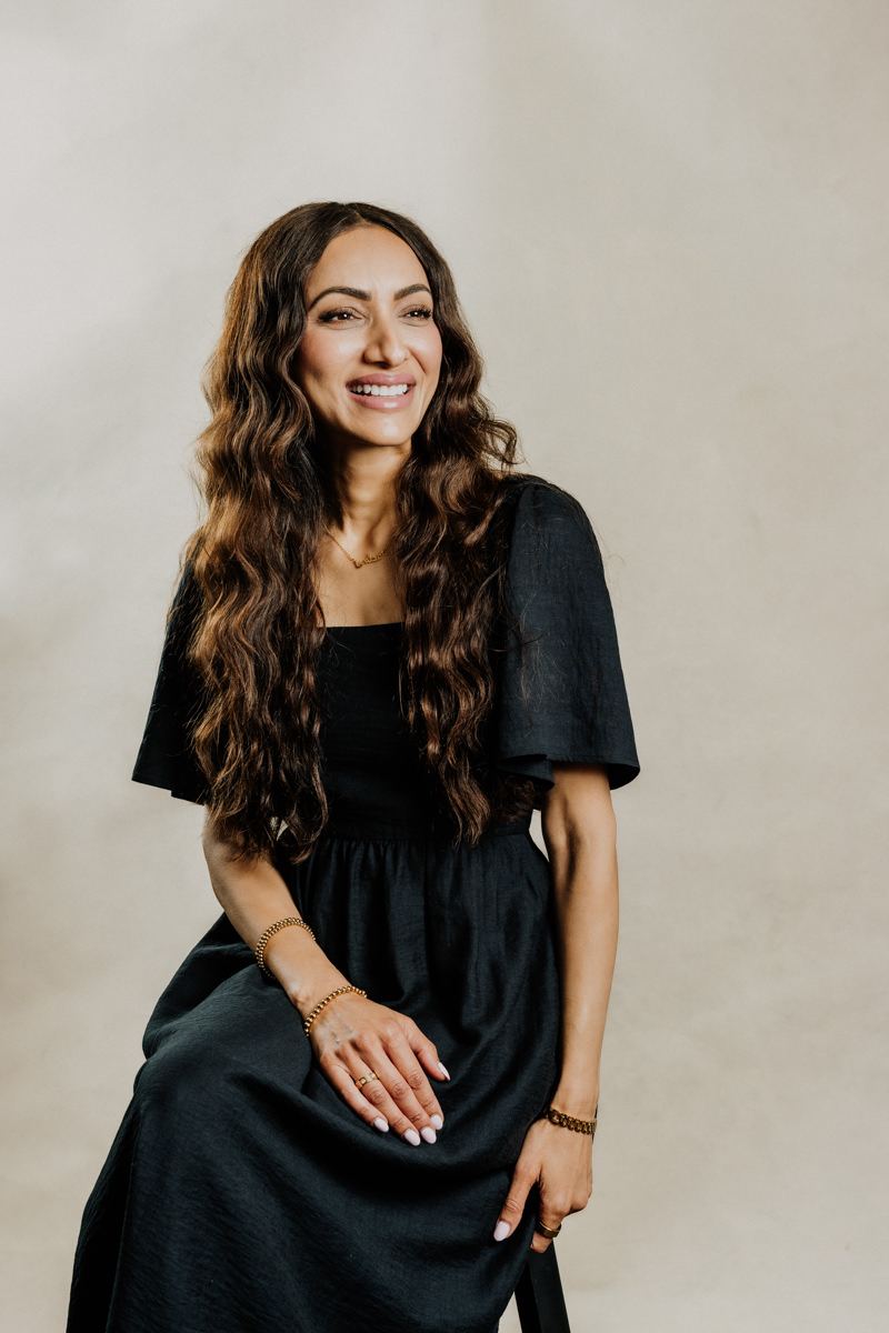 Woman in elegant editorial portrait wearing black dress and sitting on a stool.