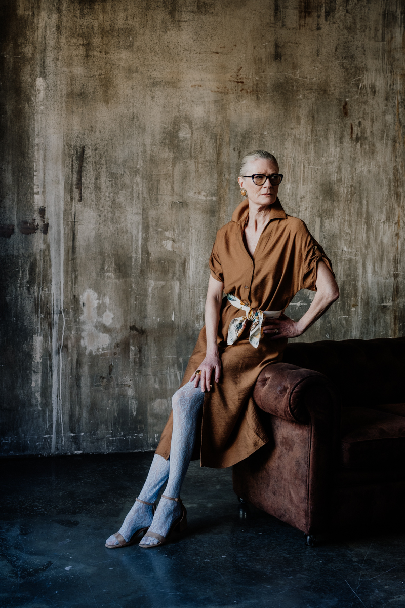 Vancouver Editorial Photographer Michele Mateus shows a portrait of older woman posed sitting on edge of sofa in rusty brown dress