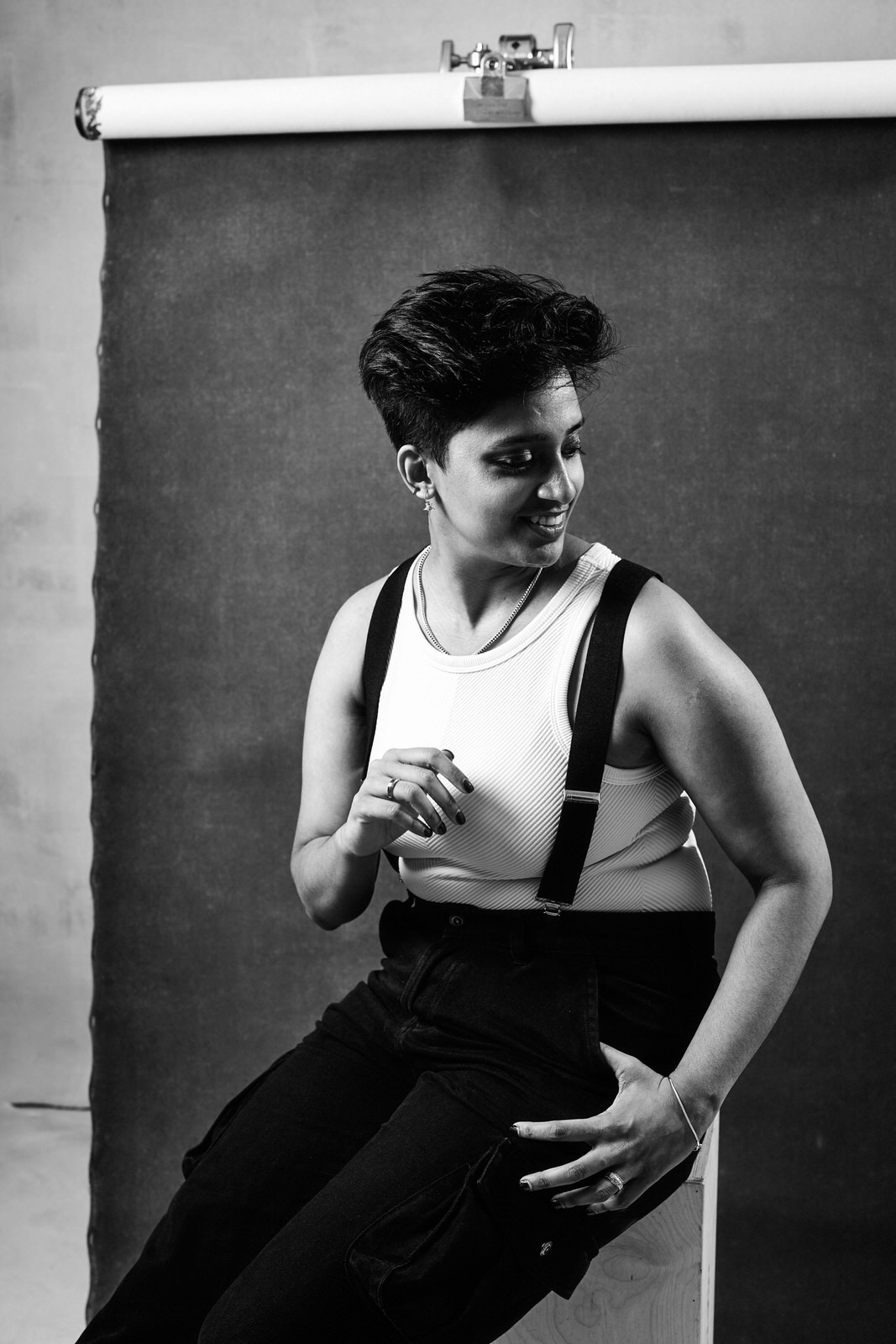 Black and white portrait of Shruti Ramani against a artistic canvas backdrop wearing a white tank top and suspenders with black pants, photographed by Michele Mateus