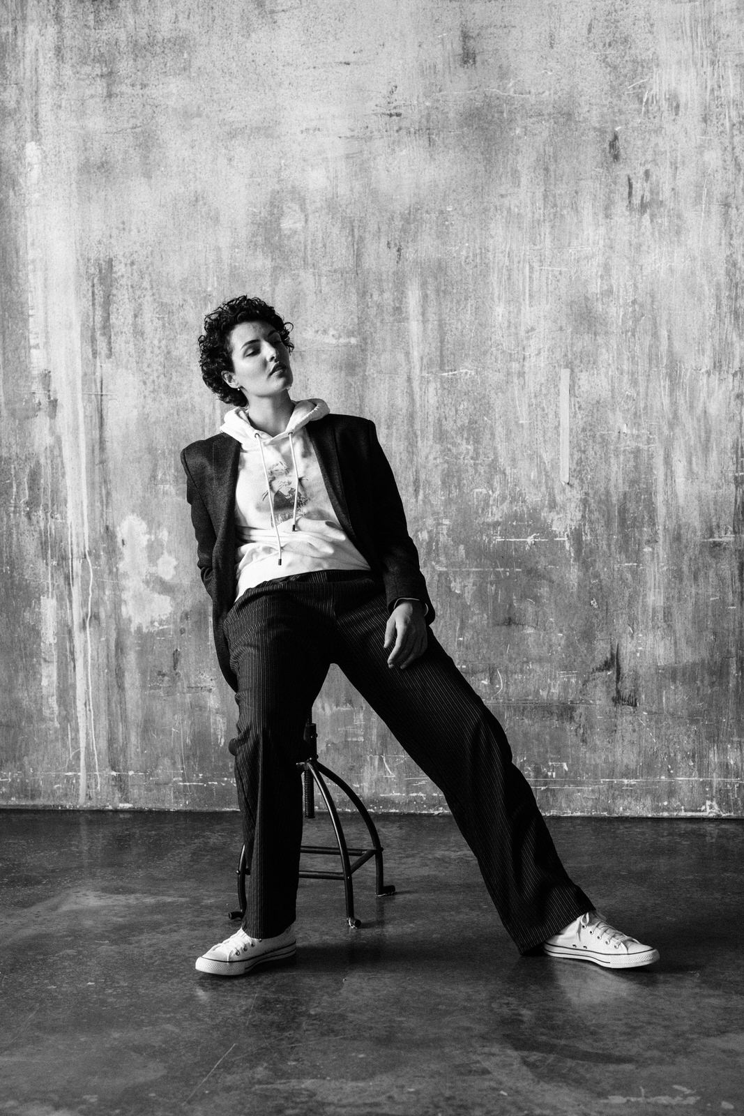 Contemporary Black and white portrait of a woman leaning against a stool with a hoodie and sweater on and suit pants and jacket against a concrete wall photographed by Michele Mateus