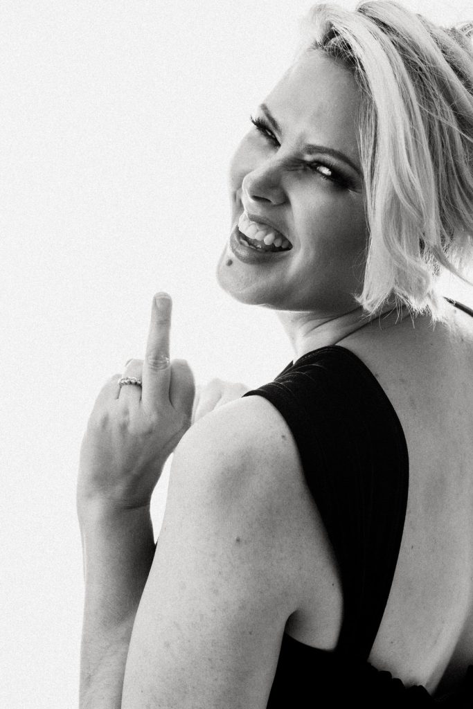 Contemporary black and white portrait of a blonde woman with short hair sticking up her middle finger in a black blazer photographed by Vancouver Portrait Photographer Michele Mateus