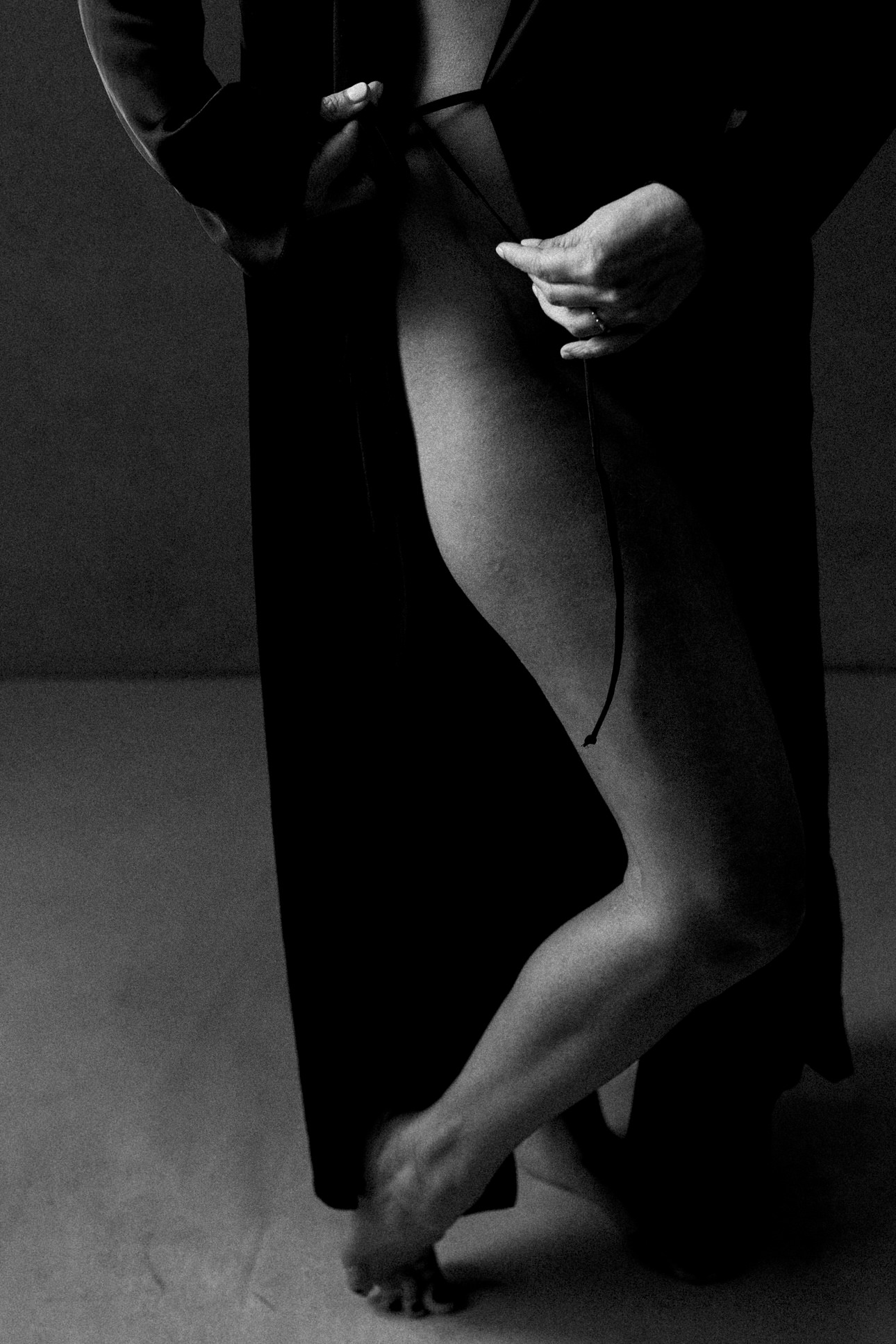Black and white boudoir portrait of woman with long black robe, her hands are tying the robe closed. The Portrait is faceless and just shows her leg.